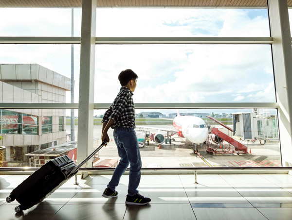 young boy pulling suitcase in airport, glass windows overlooking plane boarding